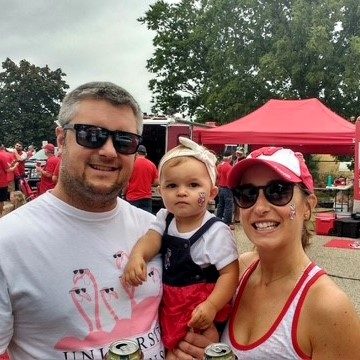 A man, woman, and toddler stand together outdoors at a football tailgate event, all wearing casual red and white clothing and sunglasses. People and red tents are visible in the background, capturing the excitement of game day.