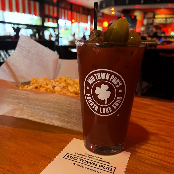A Bloody Mary with garnishes sits on a wooden table next to a bowl of popcorn in a restaurant, featuring a UW Badger football-themed coaster and the Mid Town Pub logo on the glass.