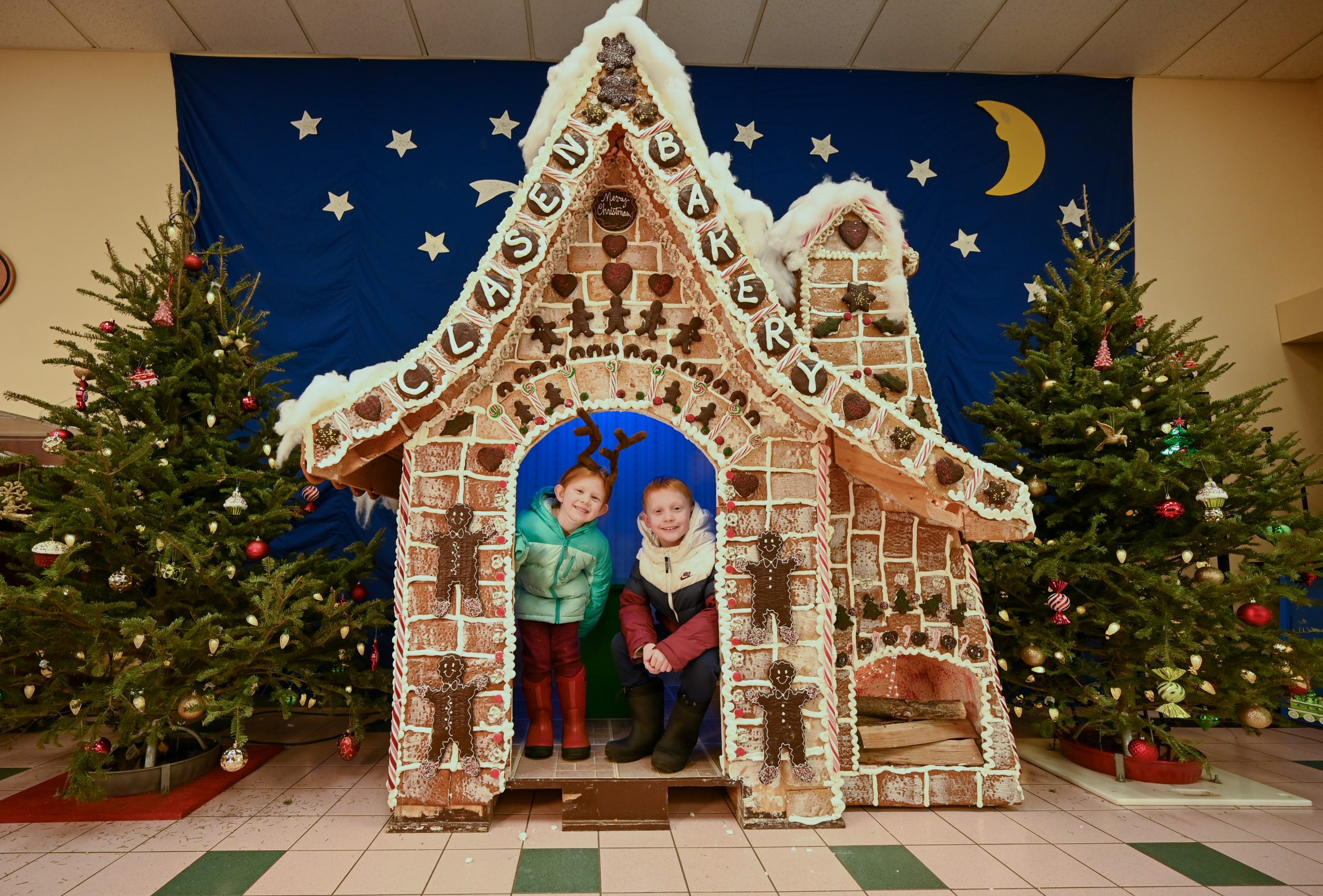 Two children stand inside a gingerbread house with a "Clasen Bakery" sign, surrounded by decorated Christmas trees and a blue backdrop with stars and a moon.