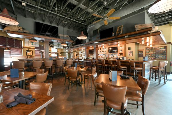 Spacious restaurant interior with brown leather chairs, wooden tables, a central bar area, and large overhead lights; no people are present.
