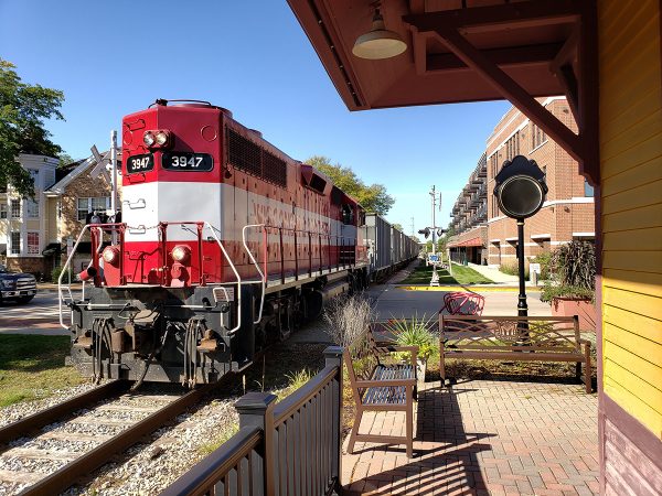 Image of red and white train passing by downtown apartment building and under the depot's eve, looking west