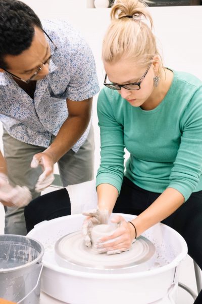 Two people engaged in pottery making; a woman in glasses shapes clay on a spinning wheel while a man watches and guides her hands.