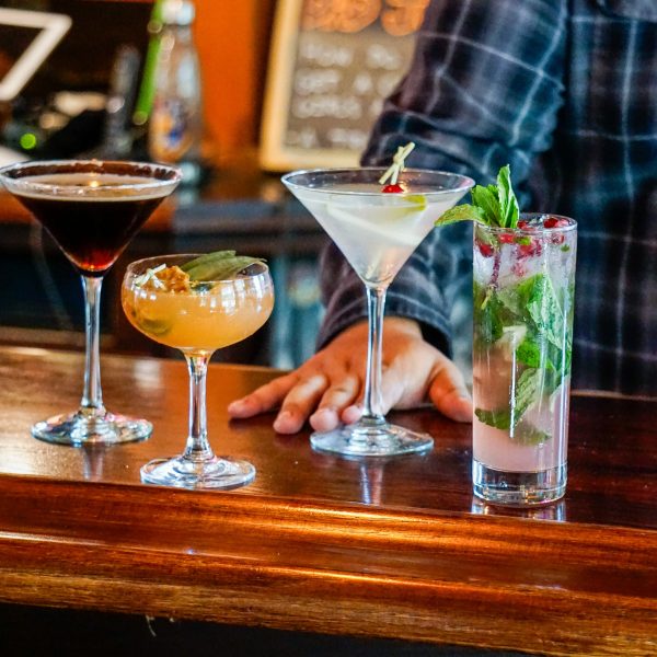A bartender is standing behind a bar with several drinks in front of him.