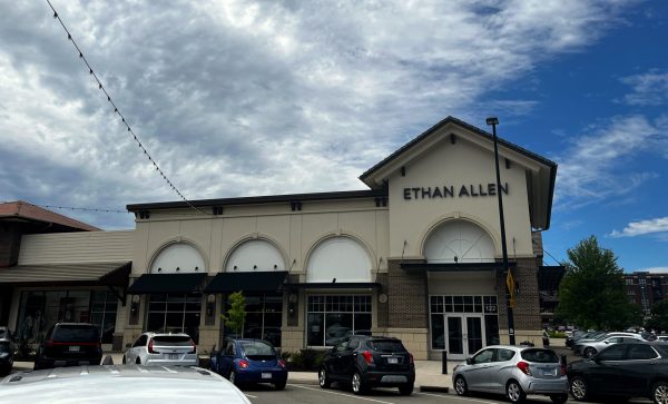 A shopping center parking lot with several cars in front of an Ethan Allen furniture store under a partly cloudy sky.