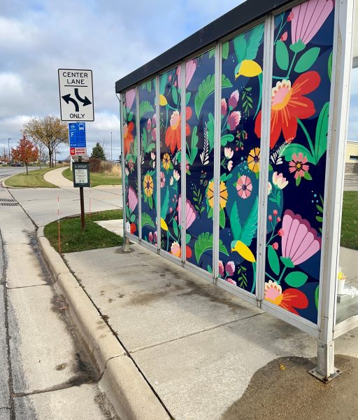 Bus stop with colorful floral mural on glass panels next to a road. Center lane sign and overcast sky in the background.