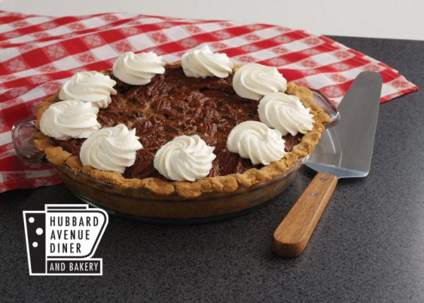 A pecan pie with whipped cream dollops on top, placed on a dark countertop with a red checkered cloth and a pie server beside it. Logo reads "Hubbard Avenue Diner and Bakery.