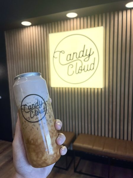 A hand holding a drink in a clear container with the "Candy Cloud" logo, in front of a wall featuring the same logo in illuminated signage.