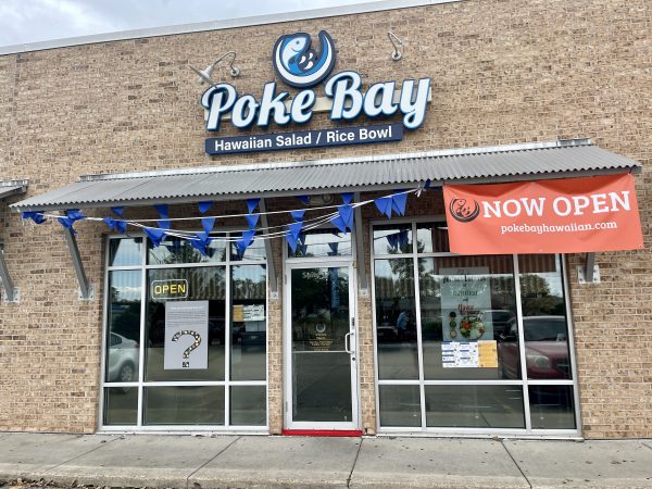 A storefront with a sign reading "Poke Bay Hawaiian Salad / Rice Bowl" and a banner stating "Now Open." The building has brick walls and glass doors and windows adorned with blue triangular flags.