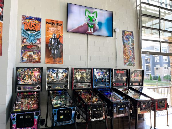 A row of six pinball machines is positioned under a wall-mounted TV displaying a green animated character. Colorful posters of "Rush," "The Mandalorian," and "Guardians of the Galaxy" are visible above.