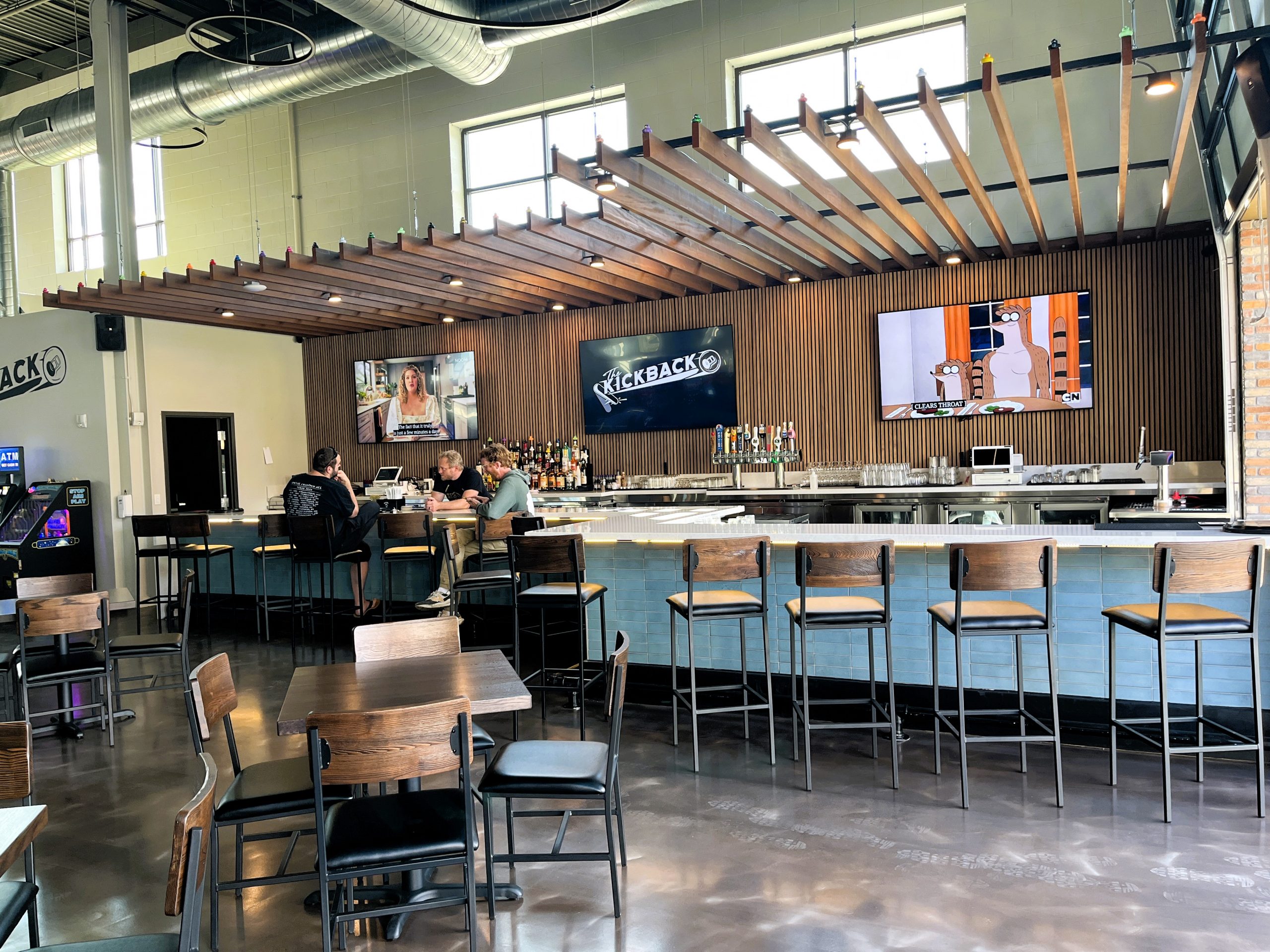 An empty modern bar with wooden furniture, a few people sitting at the counter, and TV screens on the wall. The establishment is well-lit and has a minimalistic design.