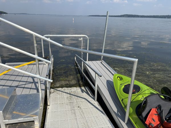 A metal dock ramp leads to a lake with a green kayak positioned on the dock; calm water and distant shoreline are visible under a clear sky.