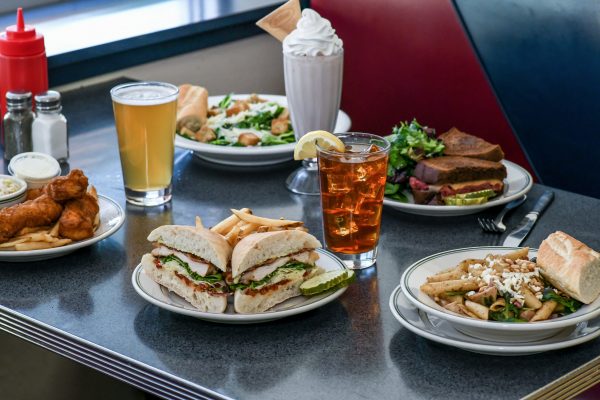 A classic diner table with plates of sandwiches, fries, salad, fish, bread, a milkshake, iced tea with lemon, a glass of beer—and a tempting slice of pie for dessert.