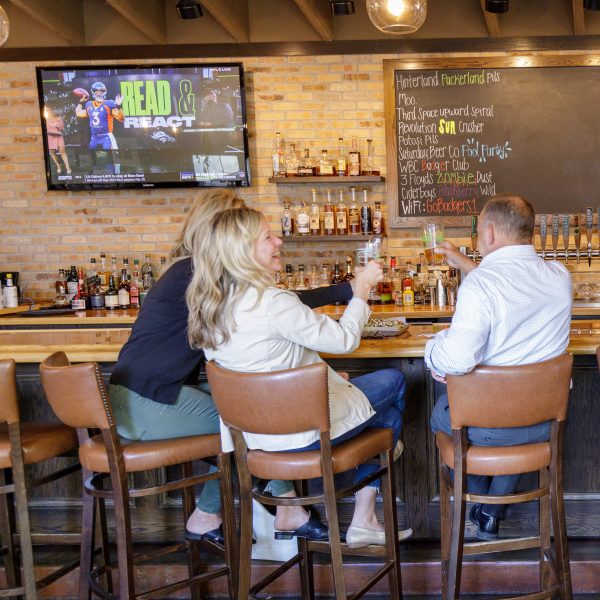 Two people sit at a bar, raising their drinks toward each other as part of their spring itinerary. A TV displays a football game above them, and a chalkboard menu is visible on the brick wall behind the bar.