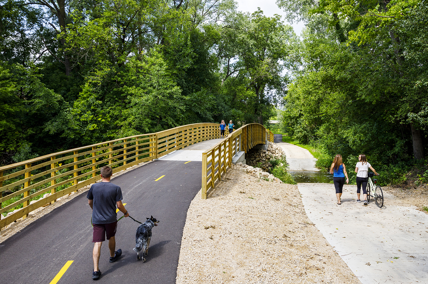 People using a multi-use trail; a man walks a dog, two women stroll, and another group approaches a wooden bridge surrounded by greenery.