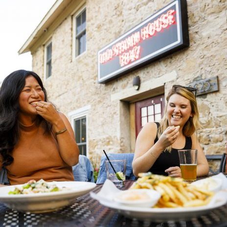 Two women enjoy patio dining at an outdoor restaurant table, smiling as they eat, with a stone building and a neon "Old Shawnee House" sign in the background.