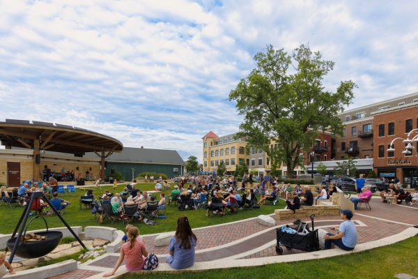A crowd of people sits on a lawn at an outdoor event near a covered stage, surrounded by brick buildings, under a partly cloudy sky.
