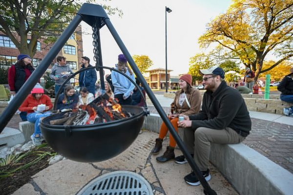 A group of people sit and stand around a fire pit outdoors at Stone Horse Green, enjoying the sunny day in this vibrant green space with trees and buildings in the background.