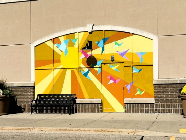 A mural featuring a large sun and colorful geometric birds decorates a building facade above a black metal bench, with two large potted plants on each side.