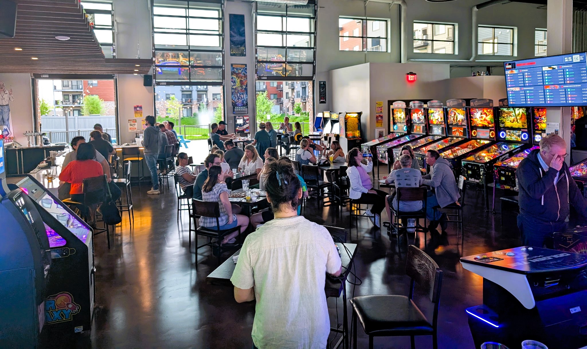 A group of people are socializing and playing pinball and arcade games inside a modern, brightly lit arcade bar.