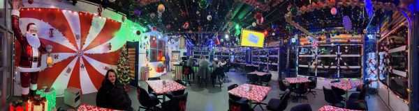 A festive indoor space with Christmas decorations, colorful lights, and tables covered in red and white cloths. People sit and stand chatting under the vibrant decor.