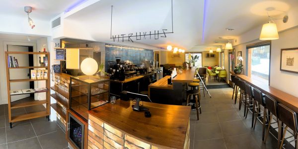Modern coffee shop interior with brick and wooden accents, bar seating, hanging lights, coffee equipment, shelves, and a plant. Sign reads "VITRUVIUS.
