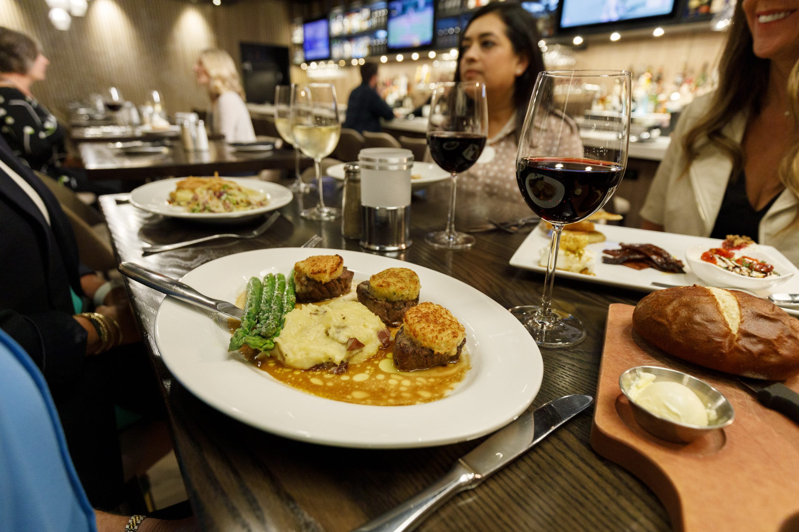 A restaurant table with diners. Close-up of a plate with three meat medallions, mashed potatoes, and asparagus. Drinks, including wine and water, are on the table. Other meals and a bread roll are visible.