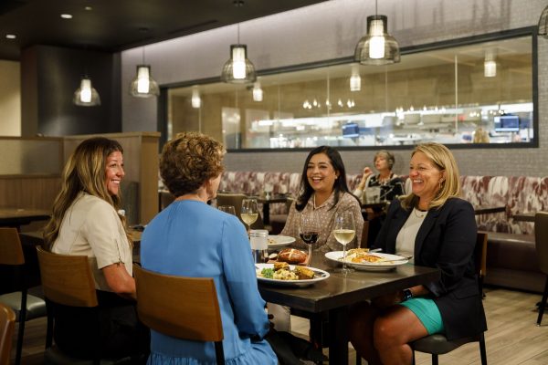 Four women sit around a table enjoying a meal and conversation in a well-lit restaurant.