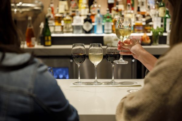 Two people are seated at a bar with four glasses of wine in front of them. One person is holding up a glass of white wine. Bottles are visible on shelves in the background.