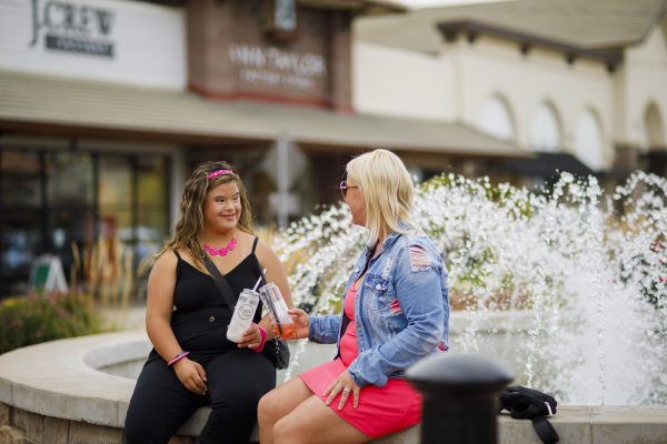 Two women sit and chat by a fountain, framed by the bustling storefront behind them. One woman is handing a bottle to the other, both immersed in their conversation as if transported to a whimsical Candy Cloud.