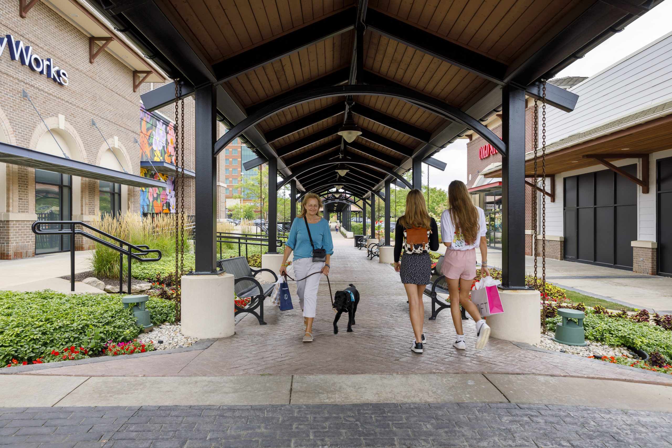 Three people and a dog walk under a covered pathway in an outdoor shopping area with stores and benches visible on either side.