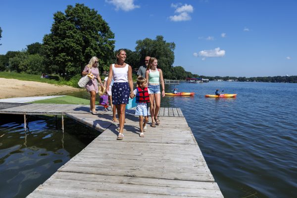 A group of people walk on a wooden dock by the lake, passing Marshall Boats as kayakers paddle on the water and trees fill the background on a sunny day.