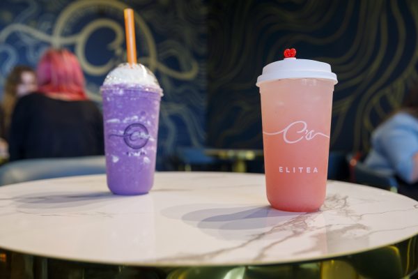 A table with two drinks: a purple beverage with whipped cream and an orange straw, and a pink drink in a branded cup with a heart-shaped lid. Blurry background with two people.