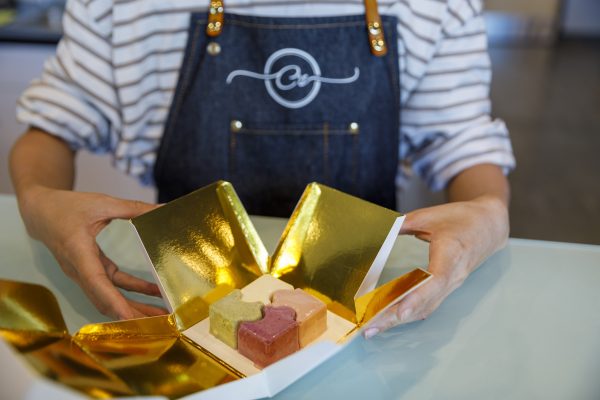 Person in a striped shirt and apron opens a golden box containing three puzzle-shaped, multi-colored desserts on a glass table.