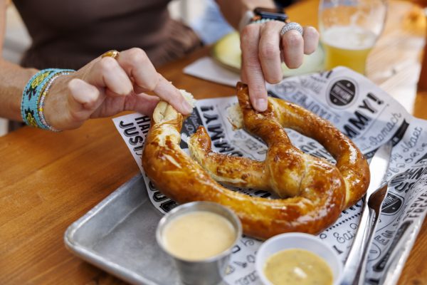 At Longtable Beer Cafe, a person breaks off a piece of a large soft pretzel on a tray with dipping sauces, food and beer nearby, sitting at a wooden table.