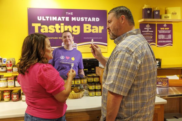 Two people sample mustard at a tasting bar while a staff member stands behind the counter at the National Mustard Museum. Jars of mustard are displayed on the counter and a sign reads "The Ultimate Mustard Tasting Bar.