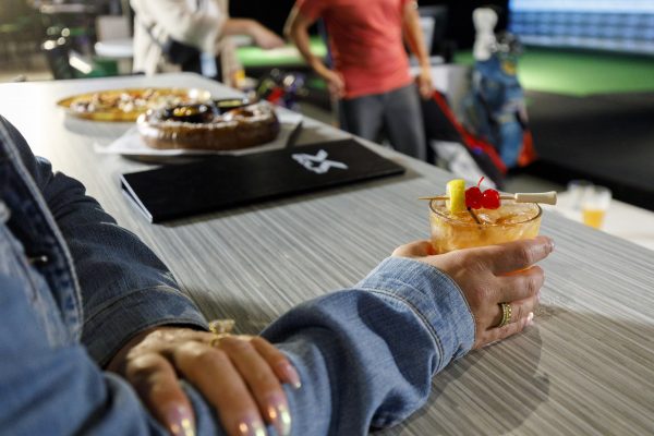 A person in a denim jacket holds a cocktail garnished with cherries and a lemon slice at a table with food and drinks, with other people and golf bags visible in the background.