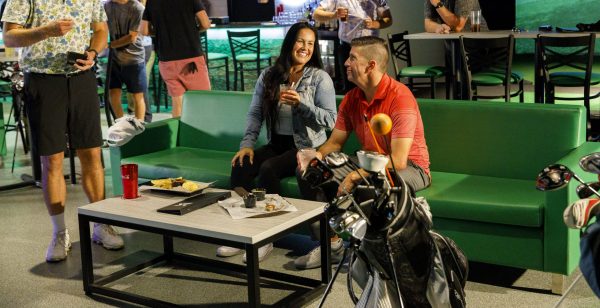 A man and woman sit on a green couch with drinks, surrounded by people. A golf bag and table with snacks are in the foreground.